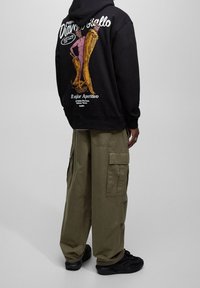 Osoba ubrana w czarną bluzę z kapturem z kolorową grafiką i napisem na plecach, oliwkowozielone spodnie bojówki oraz czarne trampki, stojąca tyłem.