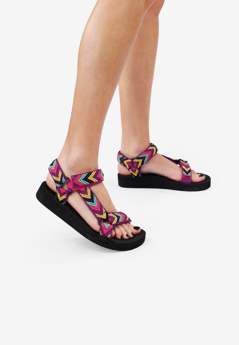Sandalias coloridas con patrón de chevron, con tiras moradas y verdes, suela gruesa negra, que cuentan con un lazo para el dedo del pie y un cierre en el tobillo.