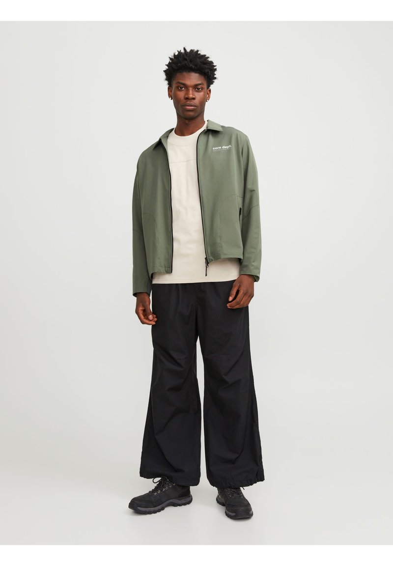 Veste verte avec un col et deux poches latérales, superposée à une chemise grise clair. Un pantalon noir ample et des baskets noires complètent la tenue.