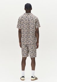 Beige Jumpsuit mit Kurzarm und Muster in Blauakzenten, mit einem kragenlosen Hals und passenden Shorts. Getragen mit einer marineblauen Kappe und Sneakers.