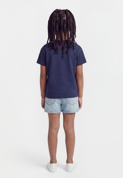 Παιδί με πλεξούδες, που φοράει μπλε σκούρο T-shirt, ανοιχτόχρωμο τζιν σορτς και λευκά παπούτσια, στέκεται με το πρόσωπο προς ένα απλό λευκό φόντο.