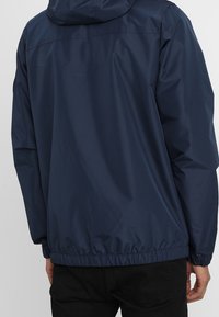 Marinblå lättviktsjacka med huva, elastisk midjeresår, slät textur och enkel design; bakifrån syns hela längden och huvens detaljer.