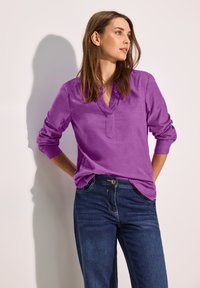 Lila långärmad skjorta med V-krage och uppvikta cuffs, bärs med mörkblå jeans. Slät textur, avslappnad passform och inga synliga mönster.