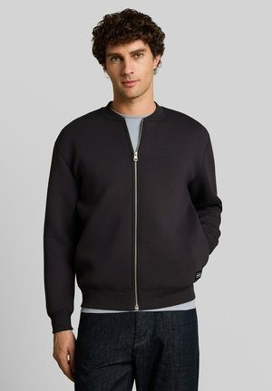 Jonge man met krullend haar die een zwart jasje met rits draagt over een lichtgrijs shirt en donkere spijkerbroek, staand tegen een effen lichte achtergrond.