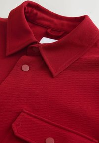 Rød bomuldsjakke med ribbet tekstur, der har en spids krave, to frontlommer og kontrasterende knapper.