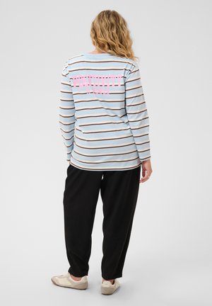 Femme aux cheveux blonds bouclés portant une chemise à rayures bleues claires à manches longues avec le texte "Positivity" dans le dos, pantalon noir et baskets beiges, debout.