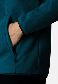 Giacca in fleece color teal con tasca con zip. Presenta una texture liscia, maniche elastiche e un cordino all'orlo per la regolazione.