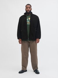 Czarna polarowa kurta na zamek z wysokim kołnierzem, noszona na zielonym t-shircie z grafiką, w połączeniu z beżowymi spodniami i czarnymi butami.