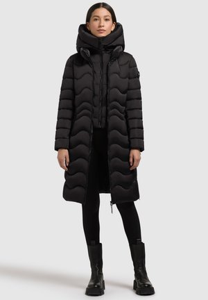 Kvinde iført en lang sort quiltet pufferjakke med høj krave, sorte leggings og sorte ankelstøvler, der står foran en ensfarvet baggrund.