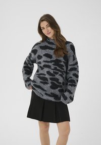 Fringe, oversize črno-sivo pulover s leopardjimi pikami, zasnova s turtleneck ovratnikom, mehka tekstura, nošen s črno ploščato krilo.