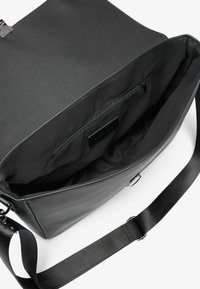 Sac messager noir en polyester et cuir, avec une poche intérieure zippée, une bandoulière réglable et un design épuré.