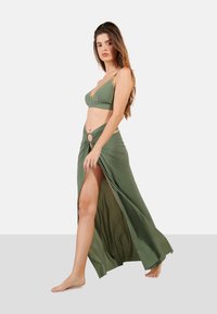 Bañador de dos piezas en verde oliva con sujetador triangular y falda de tiro alto y abertura, que presenta un acento circular en la cintura. Textura de tejido suave.