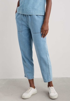 Light blue denim pants with two front pockets, relaxed fit, and ankle-length cut. Paired with white sneakers on a neutral floor background.