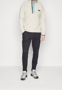 Pullover in felpa beige chiaro con colletto blu e tasca con zip, abbinato a pantaloni cargo neri con tasche laterali e scarpe sportive grigie.
