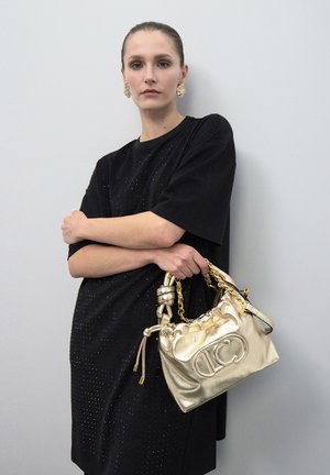 Mujer con el cabello peinado hacia atrás, lleva un vestido negro y pendientes de oro, sosteniendo un bolso dorado brillante con asa de cadena frente a una pared gris.