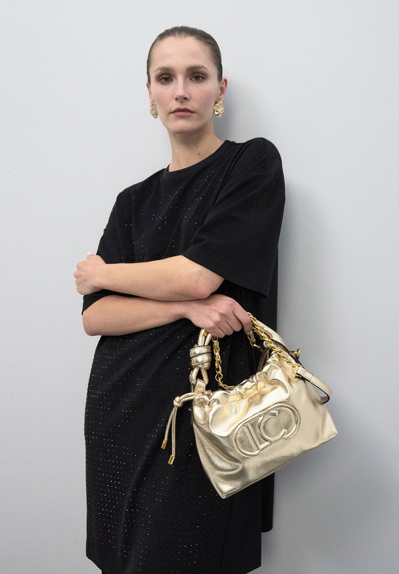 Mujer con el cabello peinado hacia atrás, lleva un vestido negro y pendientes de oro, sosteniendo un bolso dorado brillante con asa de cadena frente a una pared gris.