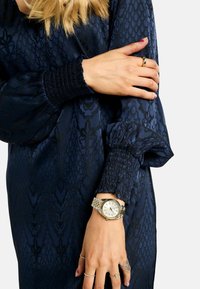 Rochie texturată bleumarin cu mâneci bufante și manșete adunate. Ceas aurit cu brățară argintie vizibil pe mână, împreună cu inele argintii.