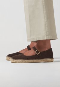 Espadrillas in suede marrone con punta rotonda, cinturino alla caviglia con fibbia e suola in corda di juta. Abbinate a pantaloni cropped beige chiaro.
