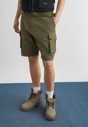 Άνδρας που φοράει cargo σορτς ελιάς, μπεζ μποτάκια με κορδόνια και λευκές κάλτσες, και μαύρο γιλέκο με ορατή πράσινη ζώνη.