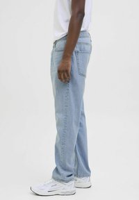 Persoană care poartă blugi drepți de culoare albastru deschis, adidași albi și un tricou alb, stând lateral pe un fundal alb simplu.