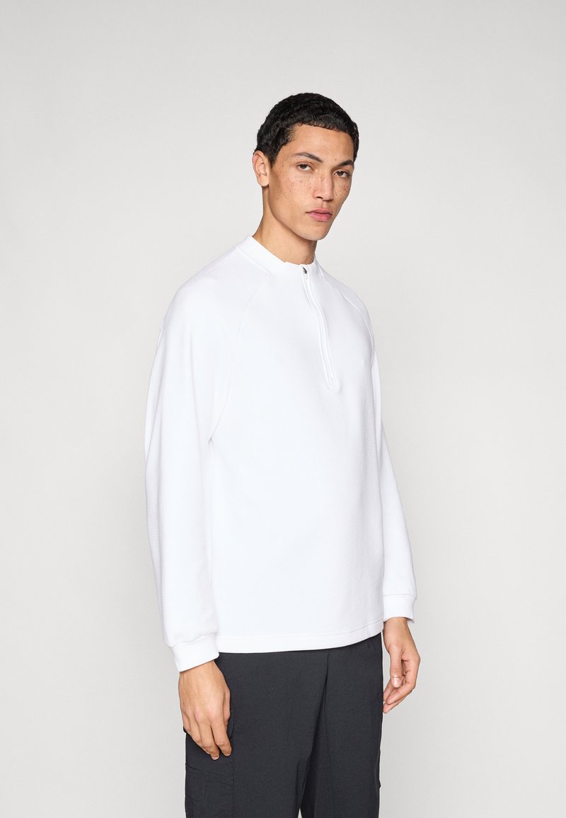 Λευκό πουλόβερ με μακριά μανίκια, ψηλό γιακά και φερμουάρ στο μισό μπροστινό μέρος. Διαθέτει υφασμάτινη υφή και ραγκλάν μανίκια. Συνδυασμένο με σκούρο παντελόνι.