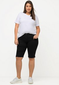 T-shirt bianca in cotone abbinata a pantaloni bermuda in denim neri. Il modello indossa sneakers bianche. Design semplice e casual con vestibilità rilassata.