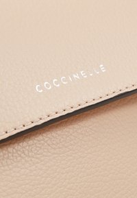 Surface en cuir beige texturé avec des coutures soignées et le nom de la marque "COCCINELLE" embossé en argent sur un rabat.