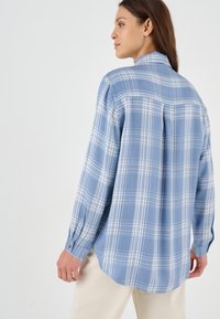 Camicia a quadri blu chiaro fatta di tessuto morbido, con colletto, polsini con bottone e orlo curvo. Presenta linee bianche e blu scure nel motivo.