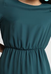 Türkises Kleid aus glattem Stoff mit rundem Ausschnitt und gerafftem Taillenbereich für eine figurbetonte Silhouette.