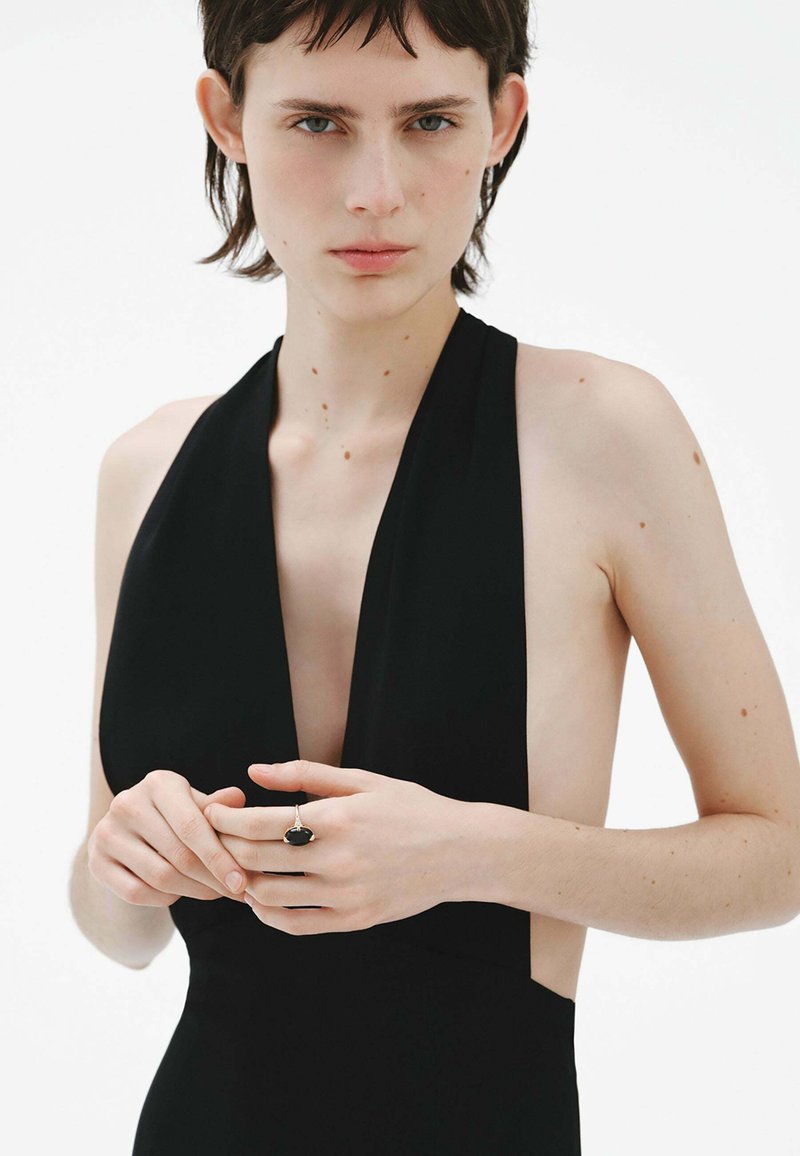Vestido negro de cuello halter con escote en forma de V profundo y recortes laterales, que presenta una textura suave. La modelo lleva un anillo con una piedra oscura en una mano.