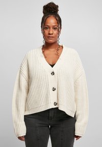 Krämfärgad stickad cardigan med V-hals, kort passform, stora svarta knappar och ribbade manschetter. Mönstrad textur och avslappnad silhuett.