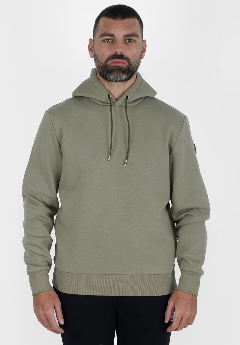 Felpa con cappuccio verde oliva in un tessuto morbido e testurizzato. Presenta stringhe di regolazione con punte metalliche e polsini e orlo a costine.