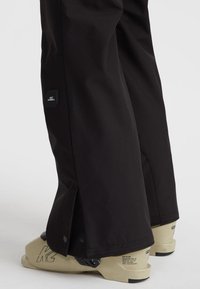 Zwarte skibroeken met drukknoopjes bij de onderkant, gemaakt van duurzaam materiaal. Logo patch zichtbaar aan de zijkant. Beige schoenen.