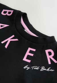 Musta collegepaita, jossa on suuret vaaleanpunaiset kirjaimet muodostaen sanan "BAKER". Alareunassa oikealla on valkoisella käsinkirjoitusfontilla pienempi teksti "By Ted Baker".