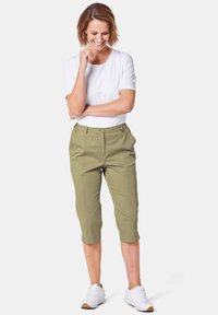 Leichtes weißes T-Shirt, kombiniert mit knielangen olivgrünen Hosen, die vorne Falten, einen Knopfverschluss und Seitentaschen haben. Trägt dazu weiße Sneaker.