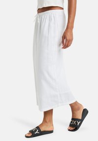 Weißes Leinen-Maxikleid mit elastischem Bund und Kordelzug, kombiniert mit schwarzen Riemchenschuhen, die ein auffälliges Logo auf dem Riemen haben.