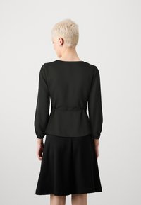 Haut péplum noir à manches longues, fabriqué en tissu lisse, avec un col rond et une taille cintrée. Assorti à une jupe noire évasée.