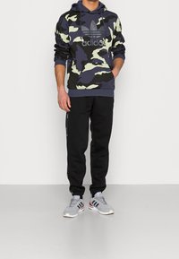 Hoodie w kamuflażowym wzorze Adidas w ciemnym niebieskim i zielonym kolorze, połączony z czarnymi spodniami dresowymi oraz szarymi sneakersami z czerwonymi i granatowymi akcentami.