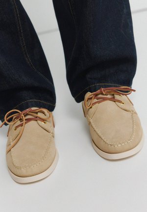 Beige Mokassin-Schuhe aus Wildleder mit braunen Schnürsenkeln, getragen zu dunkelblauen Jeans auf hellem Hintergrund.