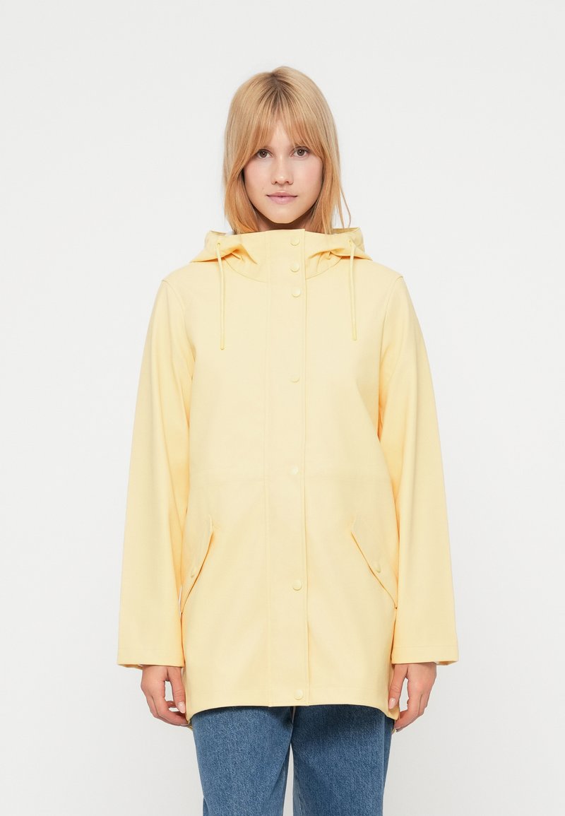 Νεαρή γυναίκα που φοράει ανοιχτό κίτρινο αδιάβροχο με κουκούλα και κουμπιά, σε συνδυασμό με μπλε τζιν, στεκόμενη μπροστά σε απλό λευκό φόντο.