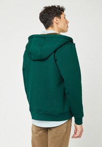 Felpa verde con una texture morbida, dotata di cappuccio, polsini a costine e chiusura con zip. Indossata sopra una camicia a quadretti azzurri chiari.