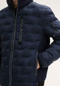 Marinefarbene gefütterte Jacke aus leichtem Material, mit gestepptem Design, doppelten Seitenreißverschlüssen und kontrastierenden dunklen Ärmeln. Verfügt über eine Reißverschluss-Brusttasche.