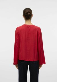 Blusa rossa con texture, maniche lunghe e scollatura rotonda. Il tessuto presenta un motivo discreto ed è abbinato a jeans in denim scuri.