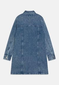 Denimjakke i mellomblå, midtlengde, med lange ermer, en knapp i mansjetten og en klassisk krage. Søm detaljene er synlige langs sømmene.