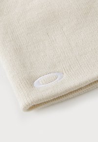 Knit beige beanie featuring a smooth texture and a white embroidered logo accent. The design is simple and seamless for comfort.