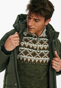 Green puffer jacket with a hood over a patterned sweater featuring diamond and zigzag designs in earthy tones. Close-up on the zipper and cuffs.