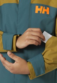 Blaue und olivgrüne Jacke mit orangefarbenem Logo, ausgestattet mit einem Reißverschluss und verstellbaren Bündchen. Eine Hand greift nach einer Karte in einer verdeckten Tasche.