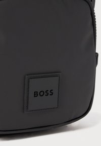 Černá nylonová taška s obdélníkovým tvarem. Texturovaný povrch s černou koženou nášivkou, na které je vyraženo jméno značky "BOSS".
