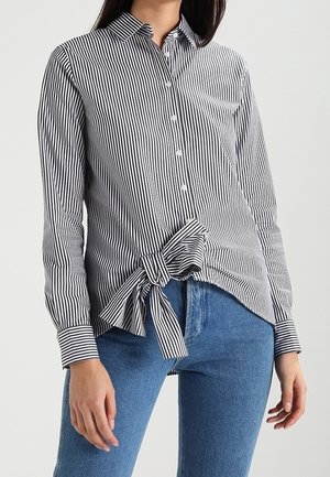 Vrouw draagt een zwart-wit gestreept overhemd met verticale knopen en een grote zijstrik, gecombineerd met een blauwe spijkerbroek, tegen een effen lichte achtergrond.