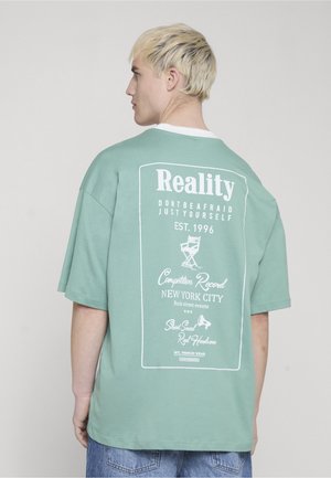 Young person with short blonde hair wearing oversized green T-shirt with white text and graphics on the back, standing against a plain light background.